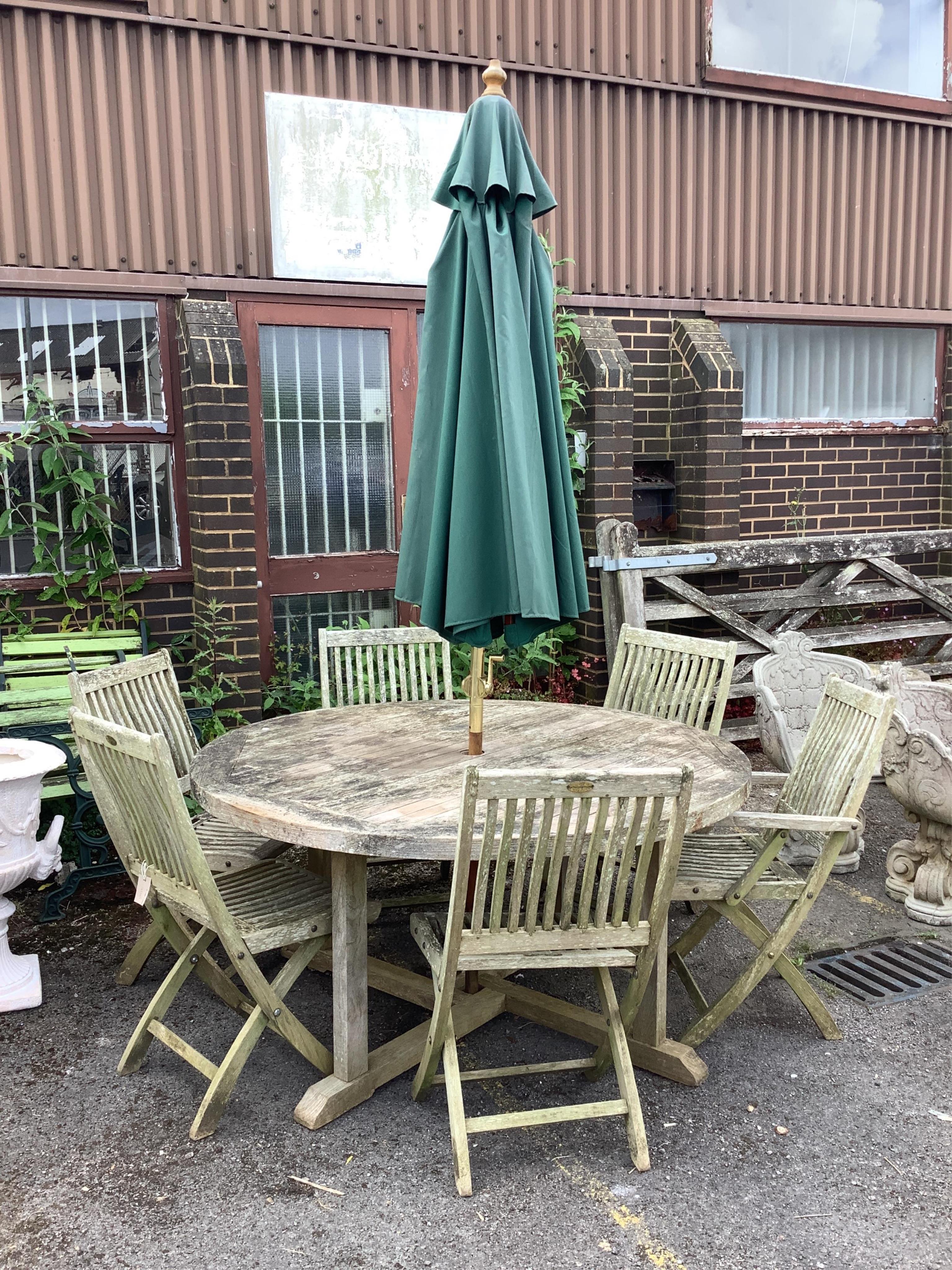 A circular weathered teak garden table, diameter 150cm, height 74cm, together with six teak folding garden chairs, two with arms and a green canvas crank parasol. Condition - fair